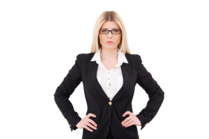 Young blonde woman wearing black glasses and a black suit, with her hands on her hips, glowers at the camera