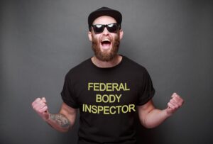 a white man with a short beard is wearing a t-shirt that says "Federal Body Inspector". He has a tattoo on his arm and is wearing a black baseball cap and sunglasses. He's got his arms out, fists up, and he's shouting, like he's cheering for something