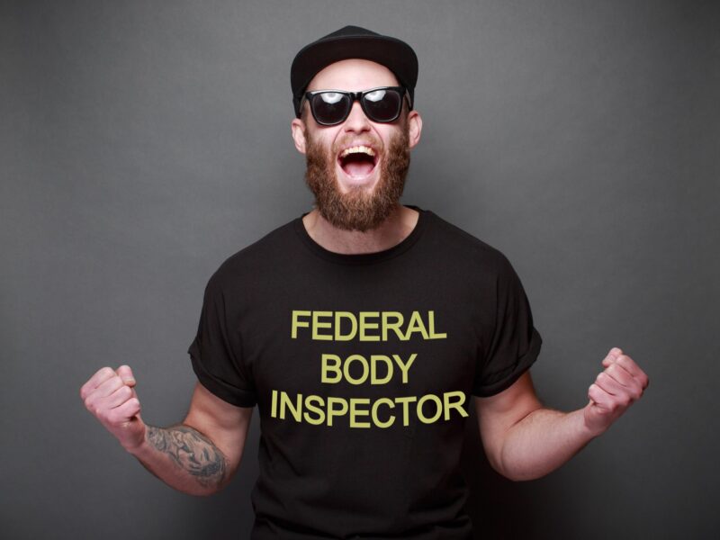 a white man with a short beard is wearing a t-shirt that says "Federal Body Inspector". He has a tattoo on his arm and is wearing a black baseball cap and sunglasses. He's got his arms out, fists up, and he's shouting, like he's cheering for something