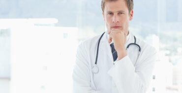 a white man in a lab coat with a stethoscope is stroking his chin in thought