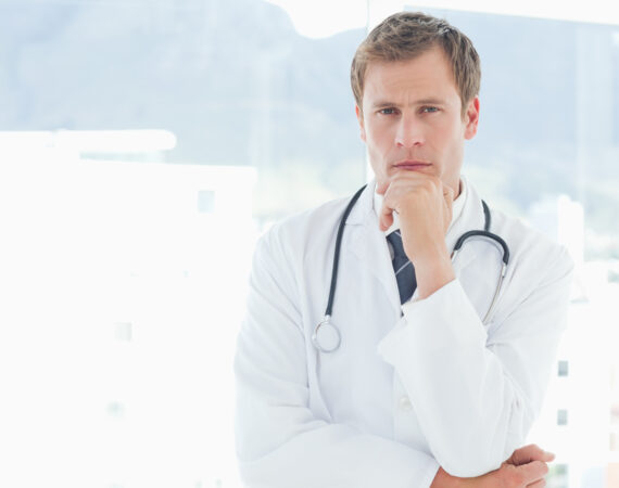 a white man in a lab coat with a stethoscope is stroking his chin in thought