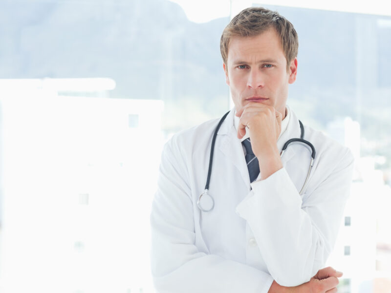 a white man in a lab coat with a stethoscope is stroking his chin in thought