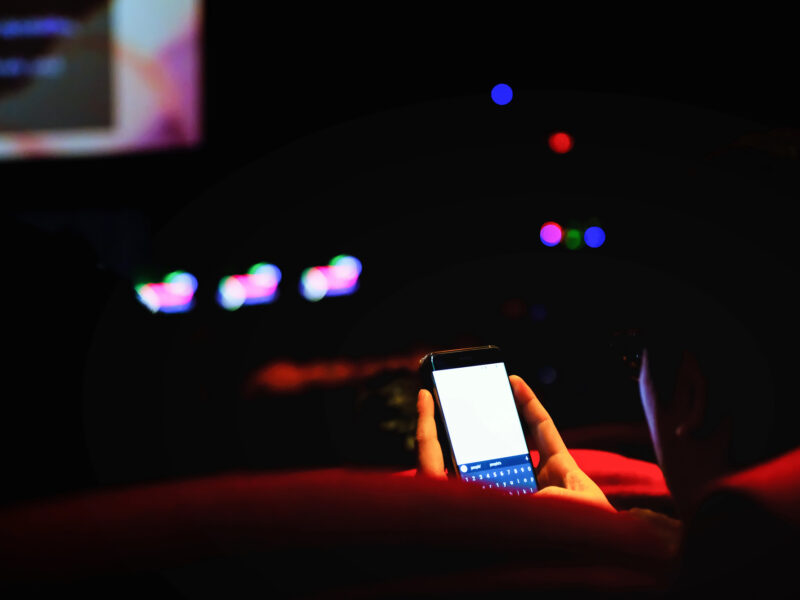 A pair of hands in a dark movie theatre are holding a very bright phone