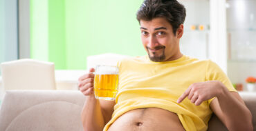 A man sitting on a couch has his t-shirt rolled up to expose his belly, which he pointing at with a happy expression
