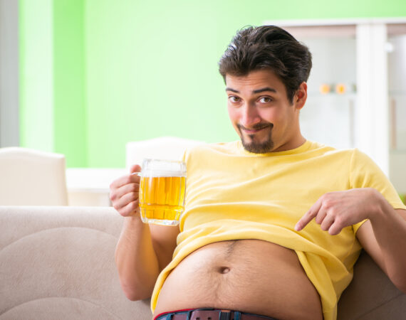 A man sitting on a couch has his t-shirt rolled up to expose his belly, which he pointing at with a happy expression
