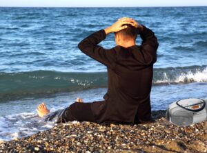 a man sitting on a beach in jeans and a black coat, his back to the camera, looking out at the ocean with his feet in the surf and his hands on his head. There's a CD boombox beside him