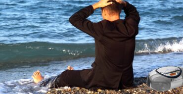 a man sitting on a beach in jeans and a black coat, his back to the camera, looking out at the ocean with his feet in the surf and his hands on his head. There's a CD boombox beside him