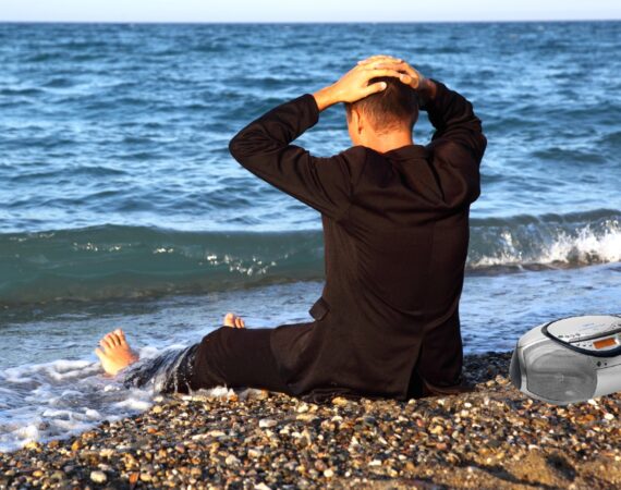 a man sitting on a beach in jeans and a black coat, his back to the camera, looking out at the ocean with his feet in the surf and his hands on his head. There's a CD boombox beside him