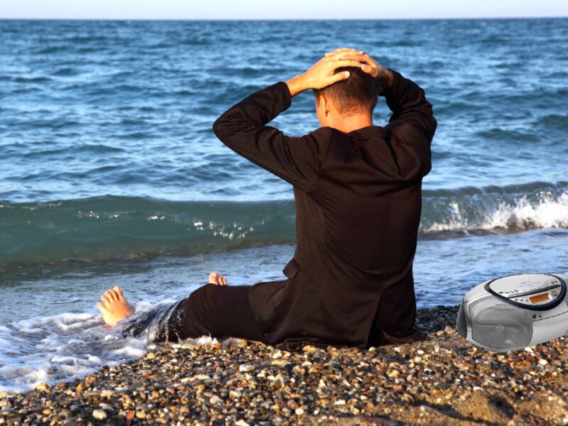 a man sitting on a beach in jeans and a black coat, his back to the camera, looking out at the ocean with his feet in the surf and his hands on his head. There's a CD boombox beside him
