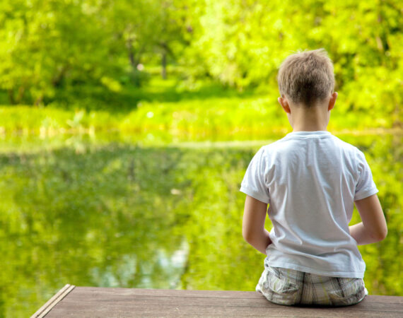 little boy sitting on dock with his back to the camera, looking at a lake surrounded by very green forest