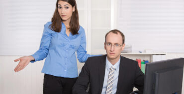 A man is looking at the camera, sitting at a desk in front of a computer, while a woman stands slightly behind him and also looks at the camera while shrugging