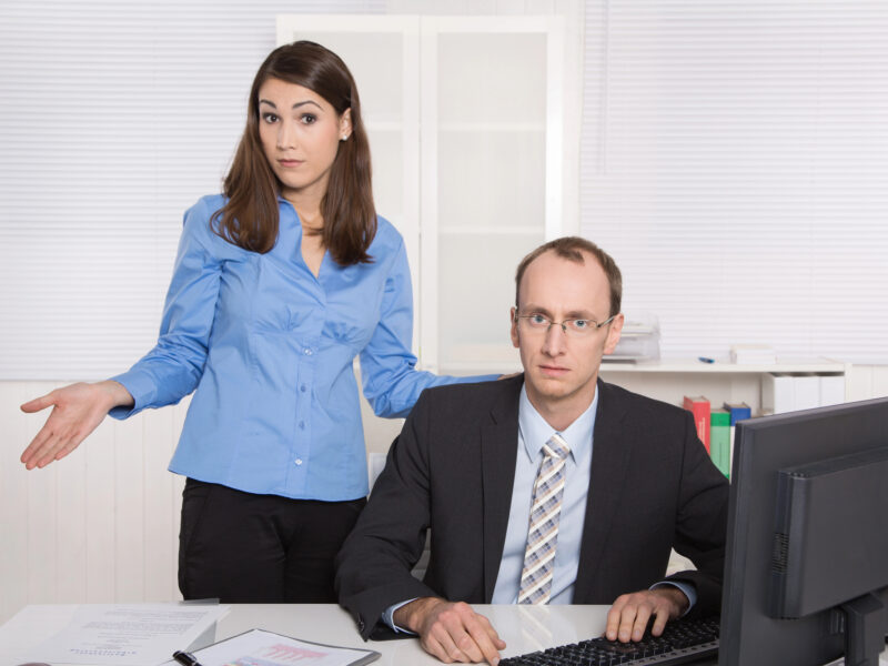 A man is looking at the camera, sitting at a desk in front of a computer, while a woman stands slightly behind him and also looks at the camera while shrugging