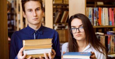 Two teenagers in a library, each holding a stack of three books and looking vaguely unhappy