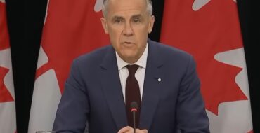 Mark Carney sitting at a desk in front of several Canadian flags
