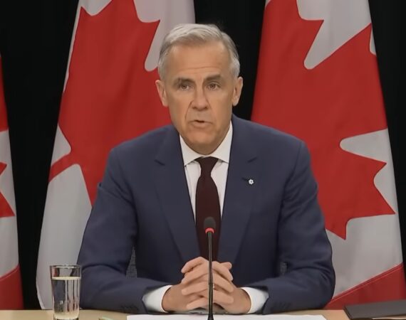 Mark Carney sitting at a desk in front of several Canadian flags